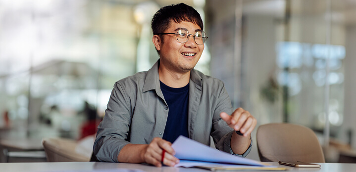 A man in a corporate setting, smiling, holding papers on a desk